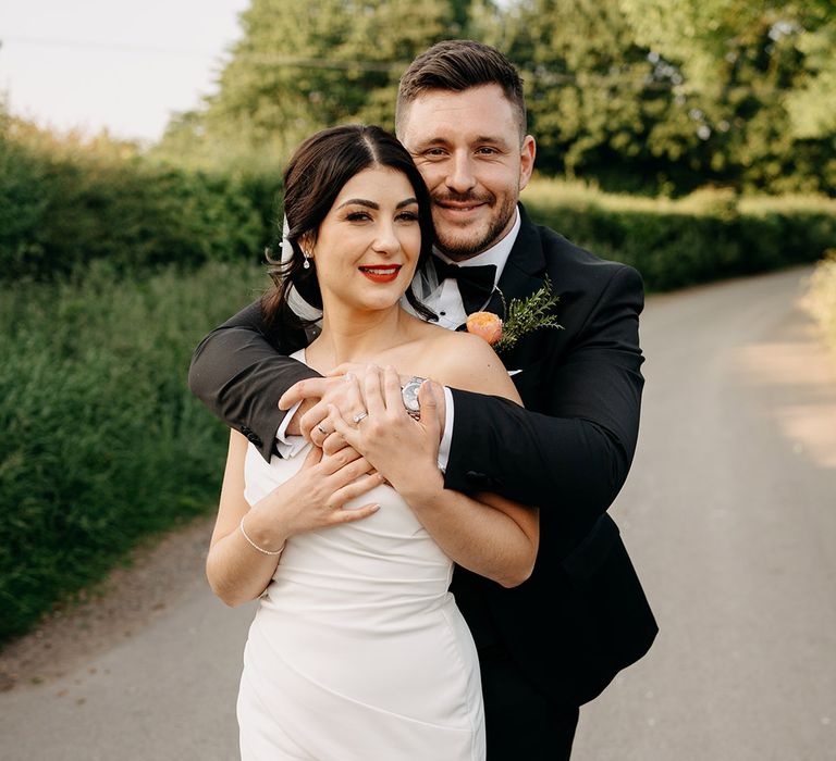 Groom with colourful buttonhole embraces the bride from behind for couple portraits