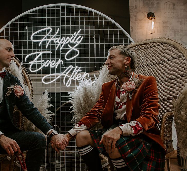 Grooms sit in front of Happily Ever After neon sign 