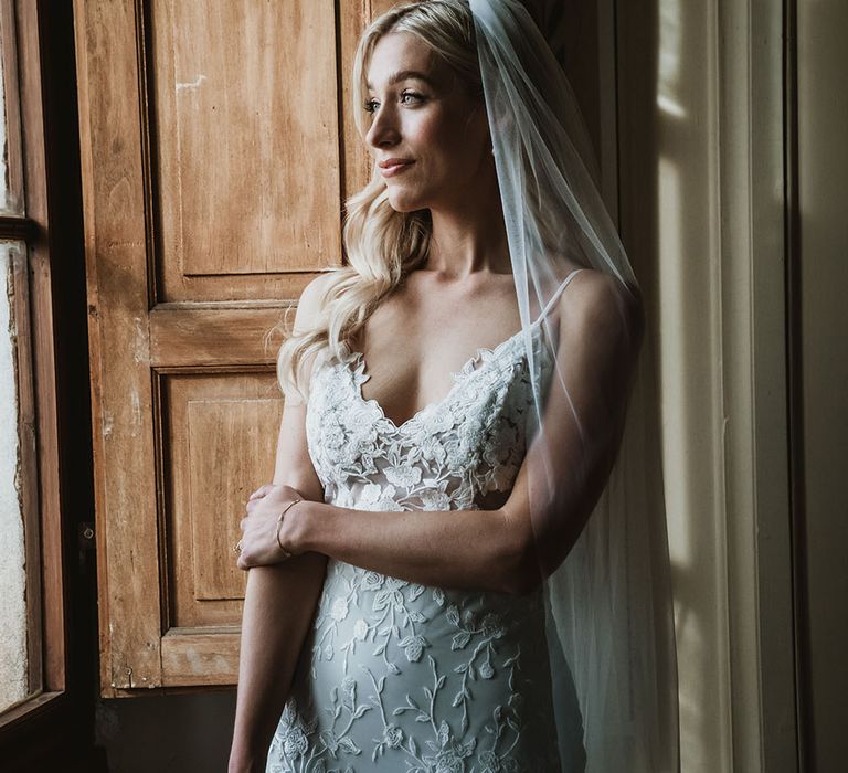 Bride looks out the window as she wears Emmy Mae Bridal lace wedding dress and Made With Love cathedral veil  