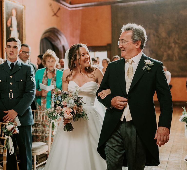 Bride holds pastel bouquet whilst wearing Suzanne Neville wedding dress and linking arms with her father who wears morning suit 