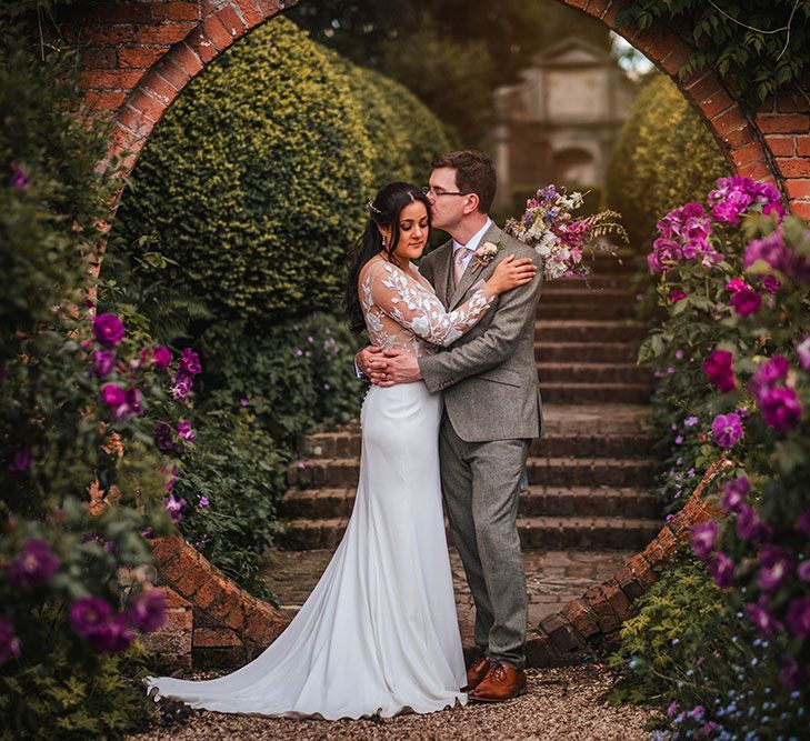 Bride & groom stand beneath circled brick archway surrounded by colourful botanicals for greenhouse wedding 