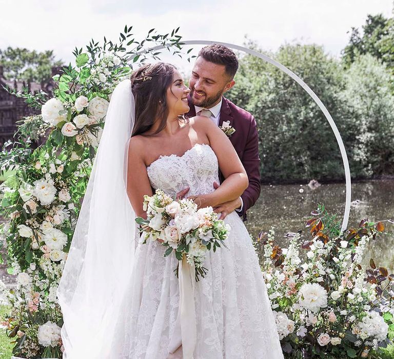 Groom in a two-piece burgundy wedding suit embracing his bride in a lace strapless wedding dress in front of a floral hoop arch 