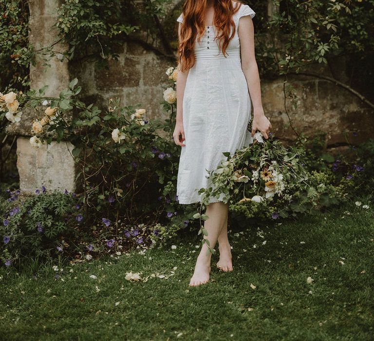 Barefoot bride with long wavy red hair in a linen midi wedding dress holding a green foliage bridal bouquet 