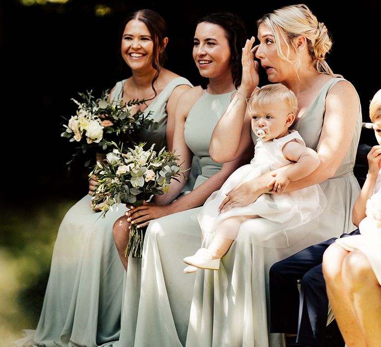 Bridesmaids in soft green dresses smile and wipe their eyes as they watch the wedding ceremony 