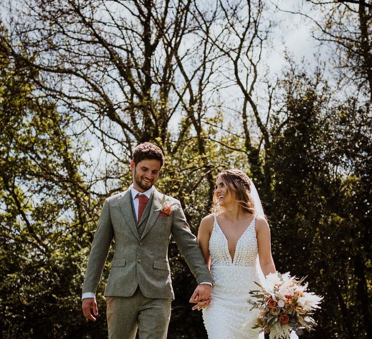 Bride in sleeveless Made With Love Bridal dress with white embellishments holding dried floral bouquet and groom in grey suit with burnt orange tie, pocket square, and boutonniere 