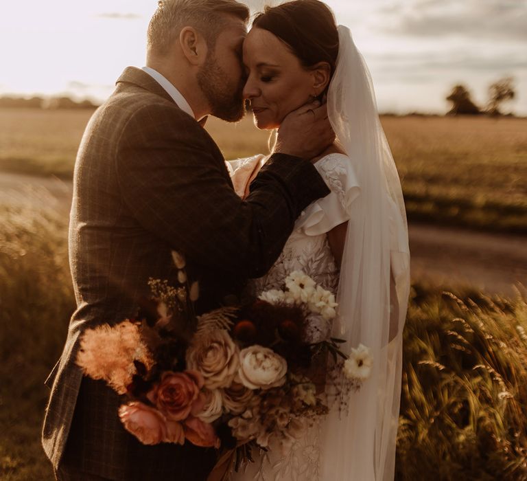 Groom in a grey checkered suit kisses the bride on the cheek during golden hour 