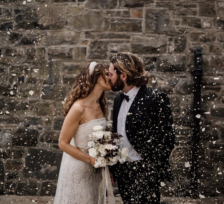 Bride in sleeveless lace wedding dress, pearl headband and black and white bouquet kissing groom in black tux