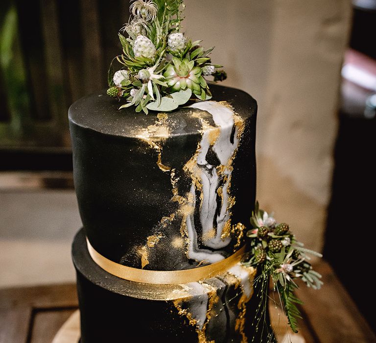 Two-tier black wedding cake with silver and golden decor and botanical wedding flowers