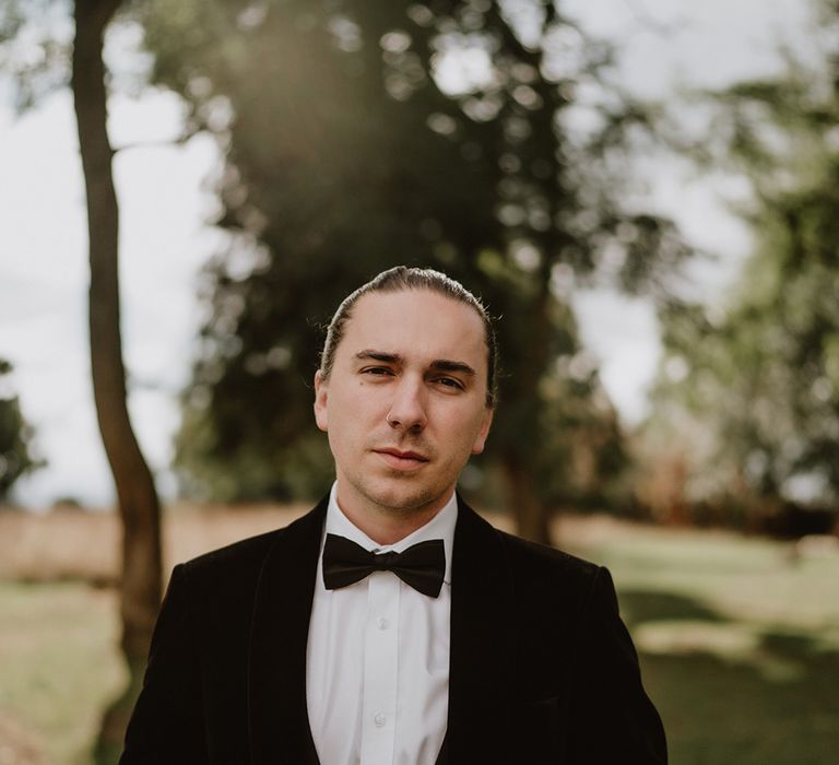 Groom in velvet look black blazer and black bowtie standing in grounds of St Tewdrics House with large statement rings 