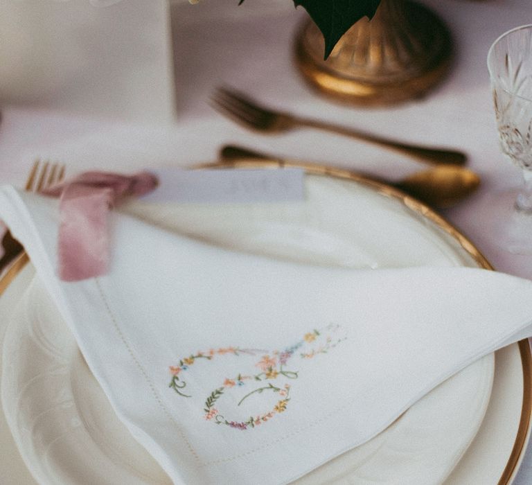 White delicate embroidered napkins with small colourful flowers and gold cutlery on neutral wedding tablescape