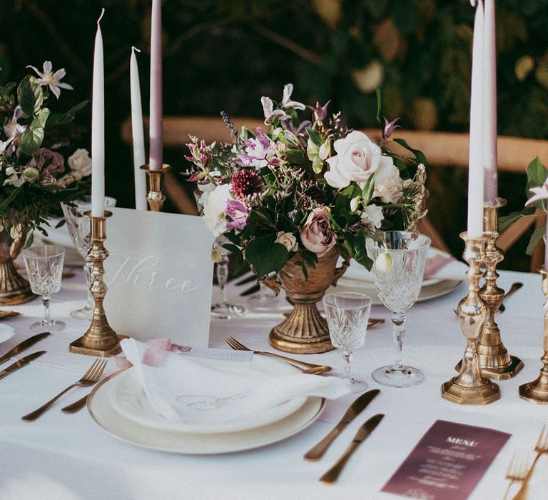 Neutral, minimalistic wedding tablescape with white tapered candles, gold candle holders, mixed floral arrangements with white carnations, purple tulips, roses and foliage, and lavender wedding menus