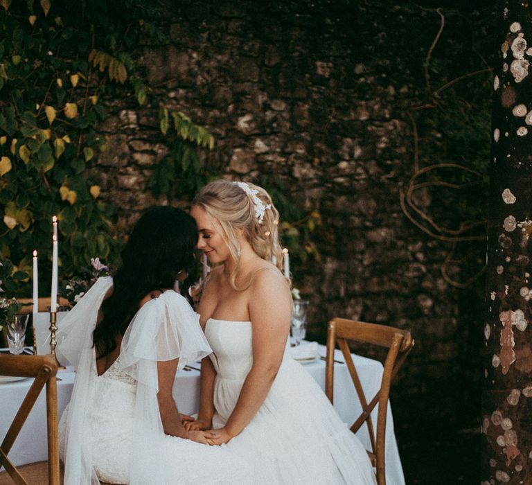 Bride in open back detail wedding dress with puff lace sleeves and bride in layered tulle sleeveless wedding dress with v neck detail holding hands at wedding reception