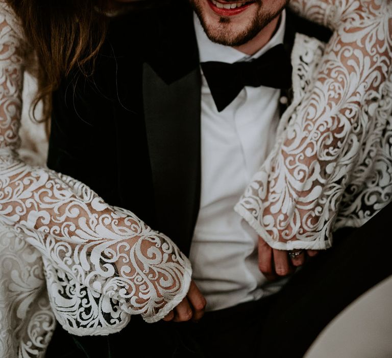 Bride in a boho laser cut lace wedding dress with bell sleeves hugs the groom from behind in a tuxedo 