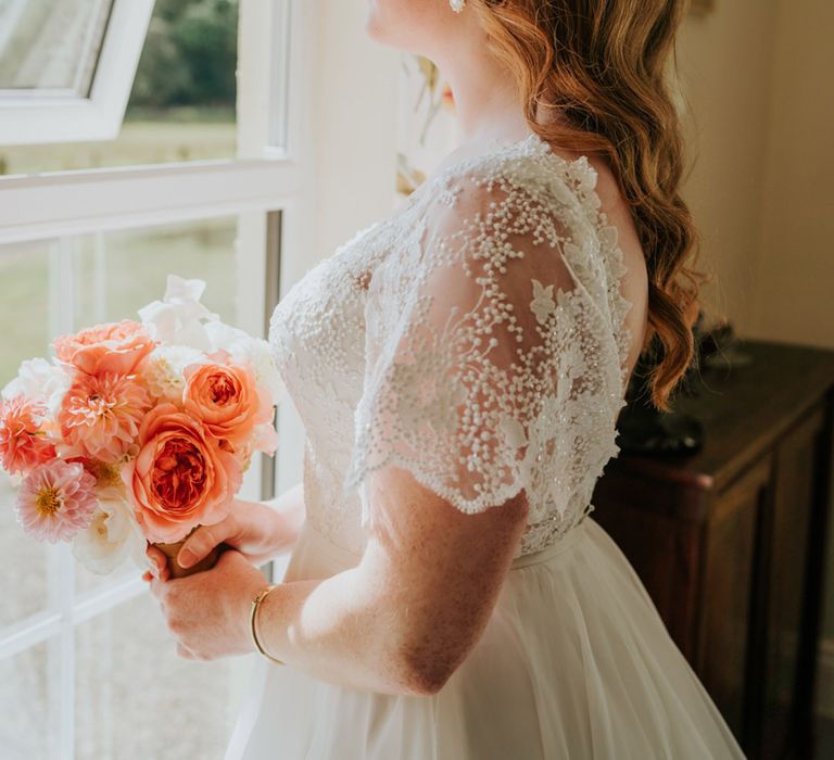 Bride wearing a lace wedding dress with a large white flower headband with pearls holding a coral and white bouquet with dahlias, peonies, and roses 