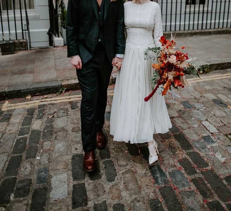 Groom in dark suit with orange tie, orange pocket square and autumnal boutonniere holding hands with bride in long sleeve lace vintage wedding dress and closed toe satin heels holding large autumnal bouquet with orange, black, green and yellow roses, peonies, foliage and pampas grass