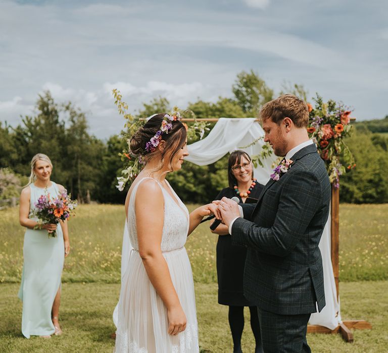 Groom places ring on his brides finger outdoors in front of wooden arch complete with white drapes and floral decor 