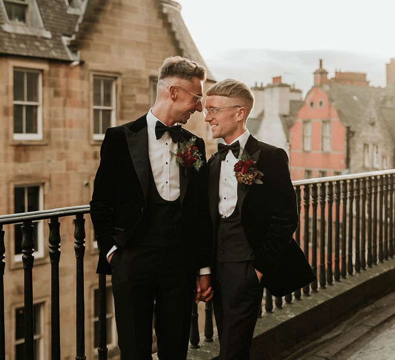 Grooms in matching black velvet tuxedos with black bowties and red rose and foliage boutonnieres embracing after stylish city wedding at The Balmoral Edinburgh