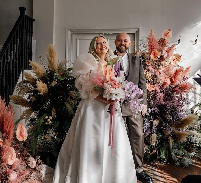 Colourful dyed pampas grass with pink, purple, and beige dried grasses and anthuriums with the bride holding a similar bouquet with orchids next to the groom 