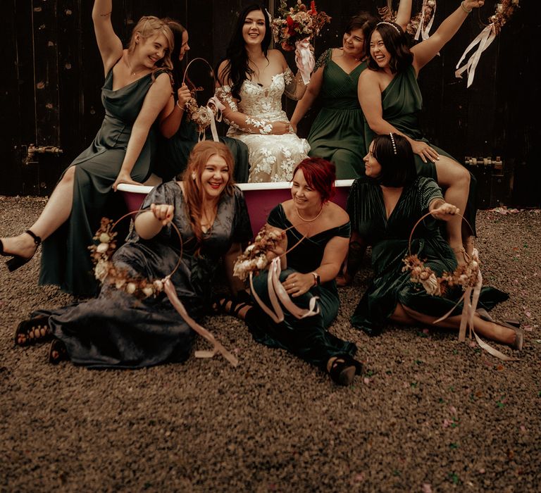 Bridesmaids in forest green mismatched bridesmaid dresses and bride in off the shoulder bodycon wedding dress with long, sheer sleeves and lace detailing sitting in copper bath at The Giraffe Shed with floral wreaths