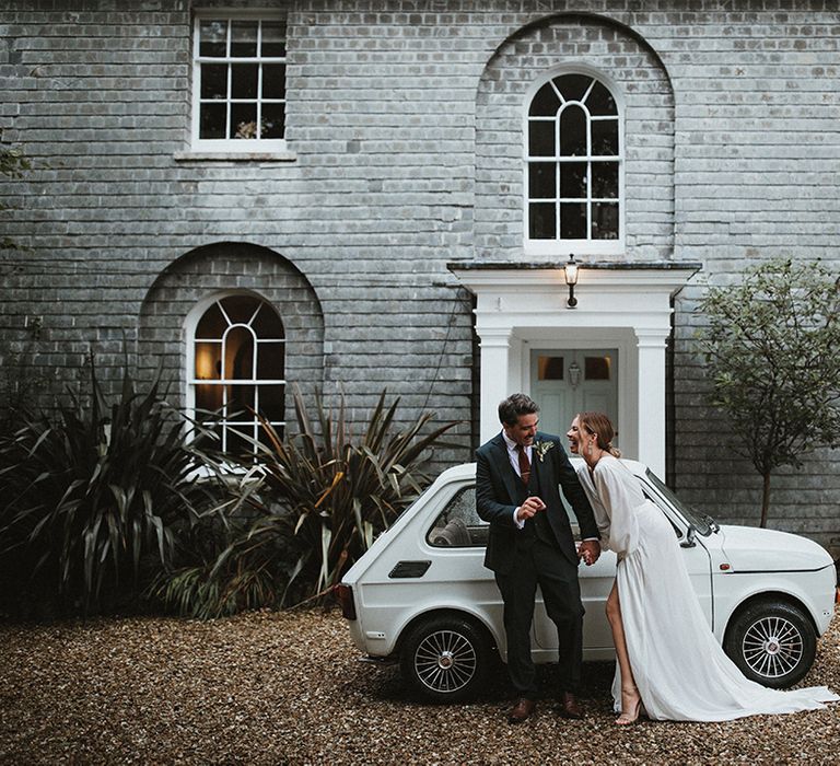 Bride in wrap styled wedding dress & groom in three piece suit stand beside vintage wedding car 