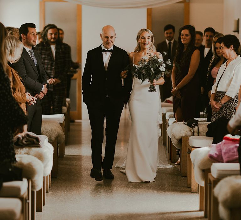 Father of the bride in black tie walks the bride down the aisle in a fitted wedding dress holding a white and green traditional bouquet with anemone 