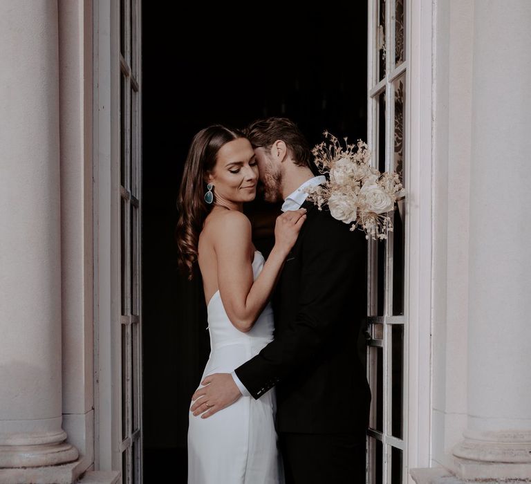 Groom in classic black blazer and suit trousers with white shirt and white pocket square and bride in strapless satin wedding dress holding bouquet with large white roses and dried flowers 