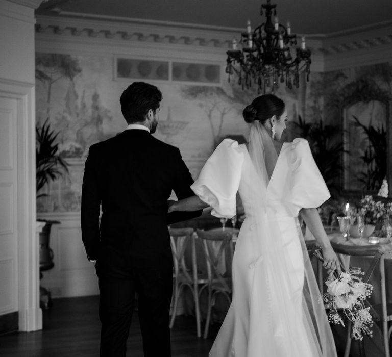 Bride in v neck puff sleeve wedding dress with church length veil and groom in black tux walking through reception room of Eaves Hall Clitheroe