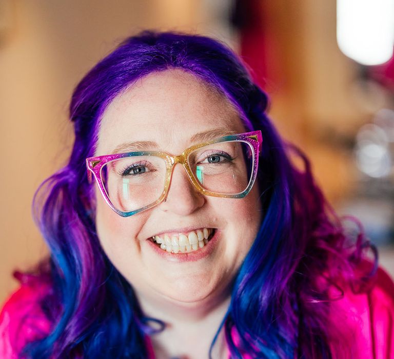 Bride with vibrant purple hair and purple cat-eye glasses wearing bright pink getting ready robe before theme park wedding 