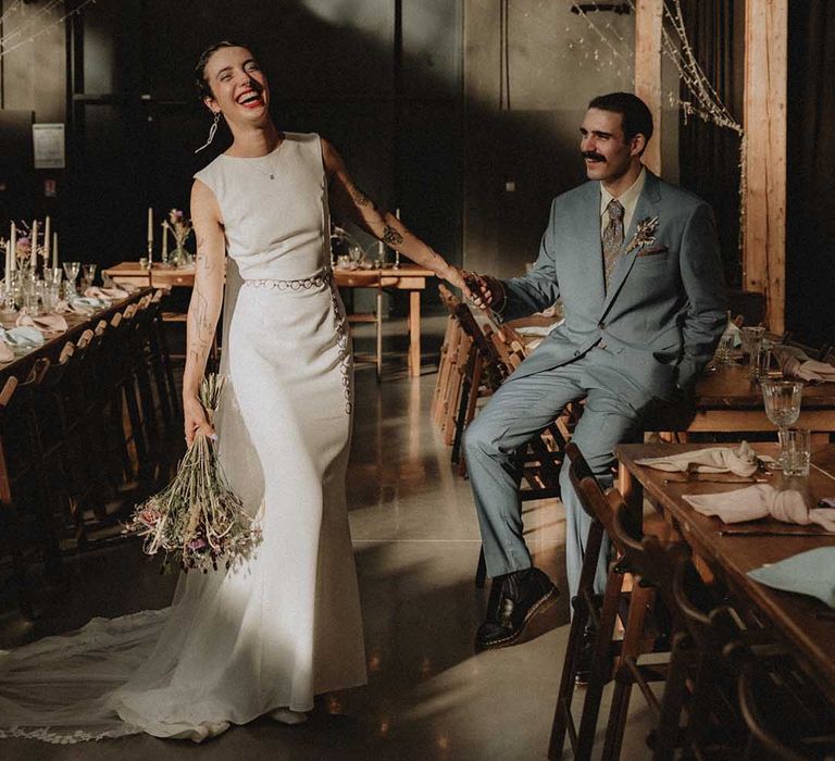 Lisa in pre-loved fitted wedding dress laughs beside Gabriel in Gucci suit as festoon lighting hangs from above 