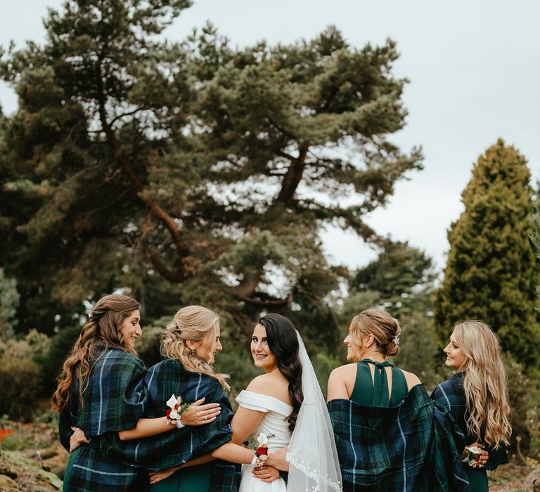 Bride looks over her shoulder as she stands alongside her bridesmaids in tartan shawl coverups and green jumpsuits 