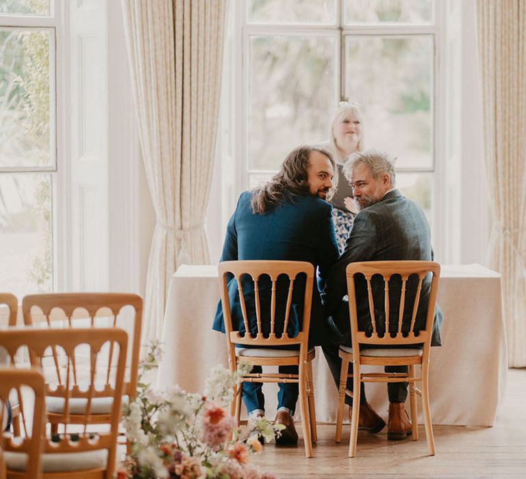 Grooms sit at table in front of window during wedding ceremony at the Cambridge Cottage 