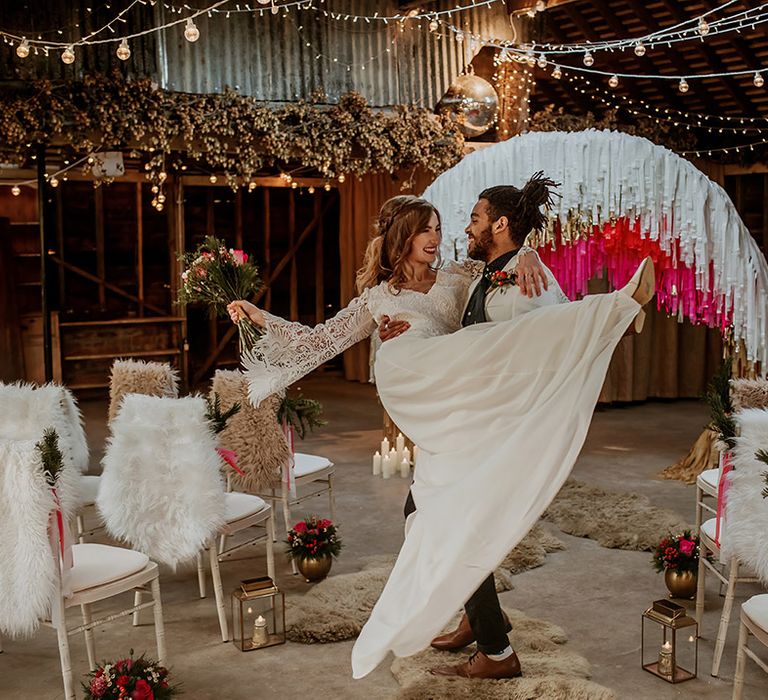 Groom in white blazer holding bride in long sleeve lace wedding dress with tassels with fairy lights, disco ball decor, aisle candles, and streamer decoration ideas 