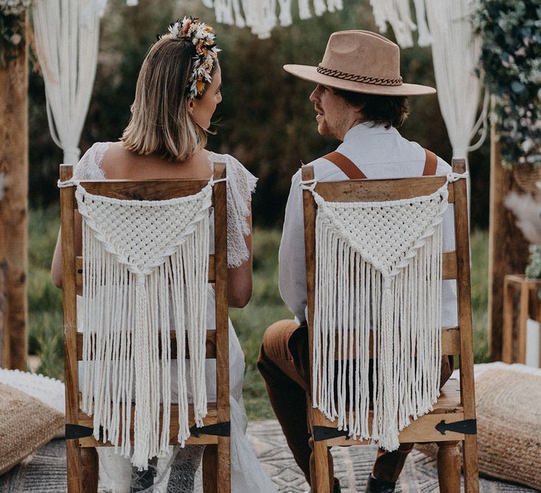 Bride in a flower crown and groom in wedding hat sit on wooden chairs for their outdoor ceremony decorated with macrame 