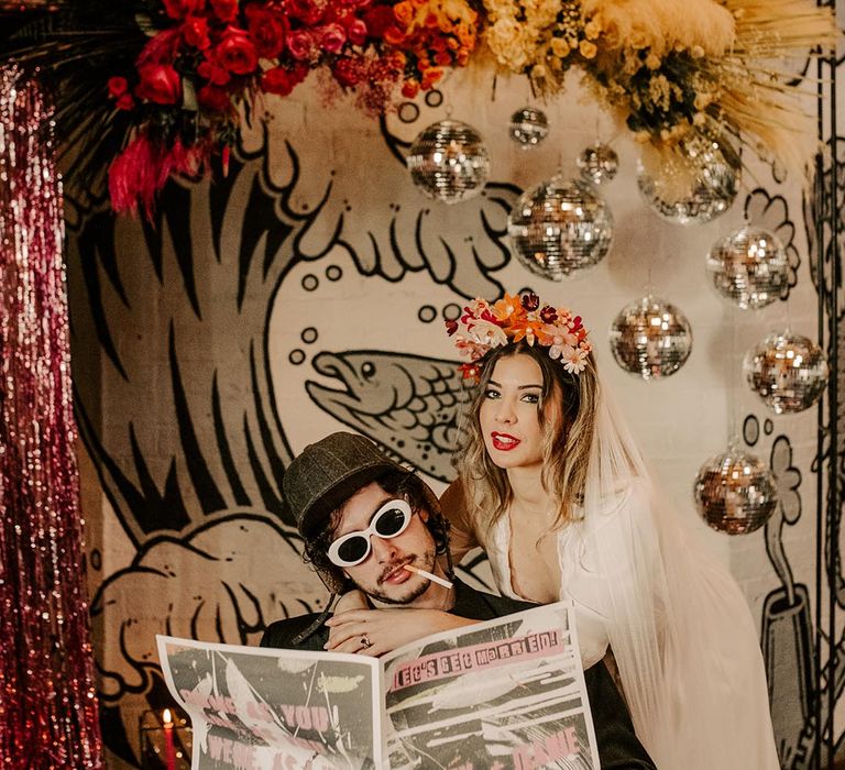 Groom in dark grey pinstripe suit, large white sunglasses and dark grey cap reading tabloid newspaper wedding stationery with bride in three quarter length sheer sleeve wedding dress with button and cinched waist details with church length veil and pink bridal flower crown sitting underneath disco ball wedding decor and pink and yellow rose flower arrangements 