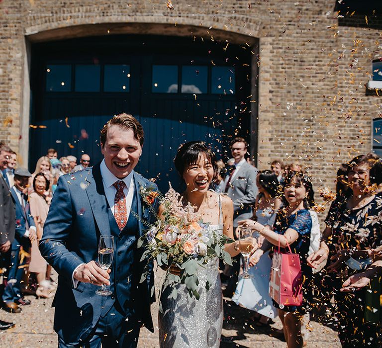 Bride in silver wedding dress walks through colourful confetti exit with her groom outdoors 