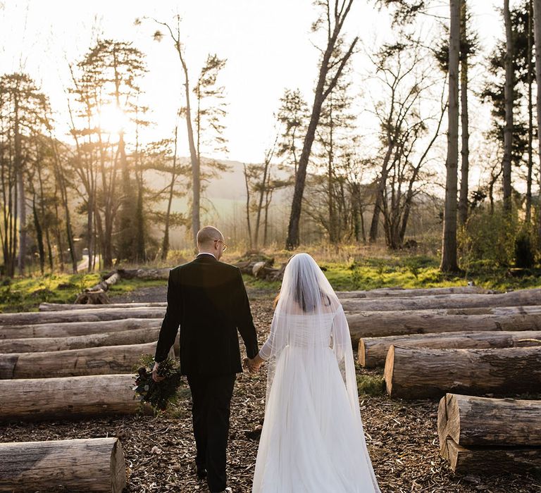 Bride in a tulle length wedding dress and cathedral length veil walks with the groom during the sunset 