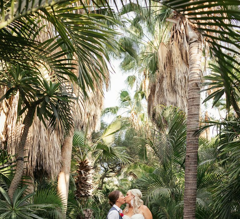 Brides kiss outdoors surrounded by palm trees during outdoor wedding in Mexico