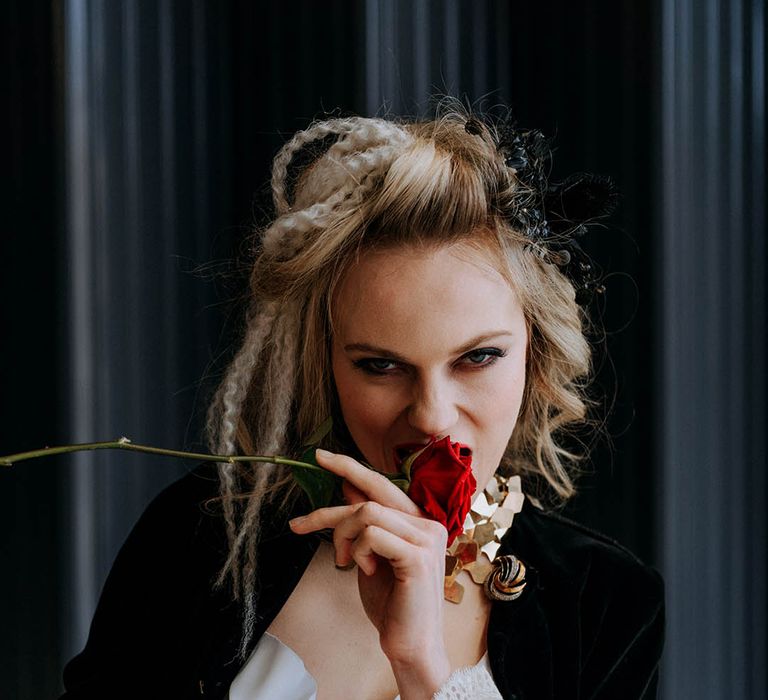 Bride in sculpted bandeau maxi wedding dress with black fitted jacket with cream lace sleeves, crimped hair and black artistic hair piece biting into red rose