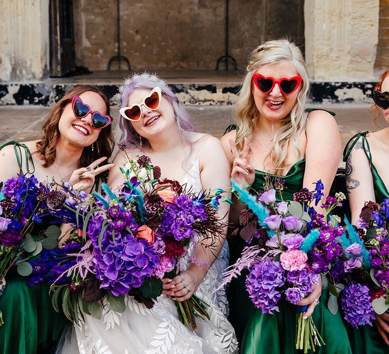 Bride in appliqué bridal gown and cathedral length veil and white heart shaped wedding sunglasses and holding bright purple bridal bouquet with purple hydrangeas, pink garden roses, purple orchids, lilac carnations, dried flowers and peacock blue feathers sitting with bridesmaids in satin forest green bridesmaid dresses and heart shaped sunglasses 