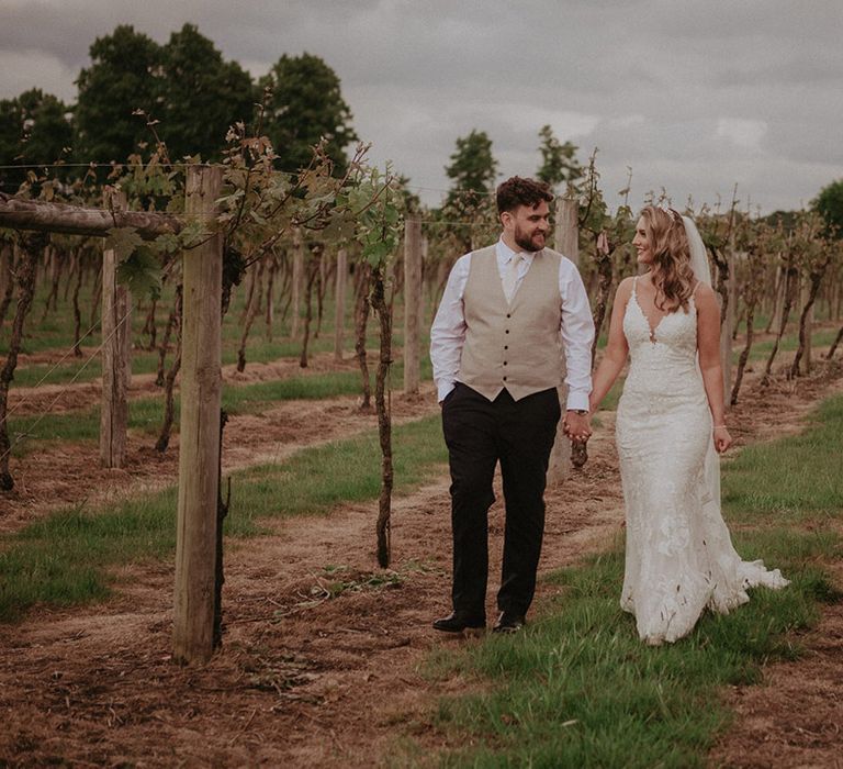 The groom wears a pale beige waistcoat and white shirt with dark trousers alike hand in hand with the bride in a fitted lace wedding dress and tiara 