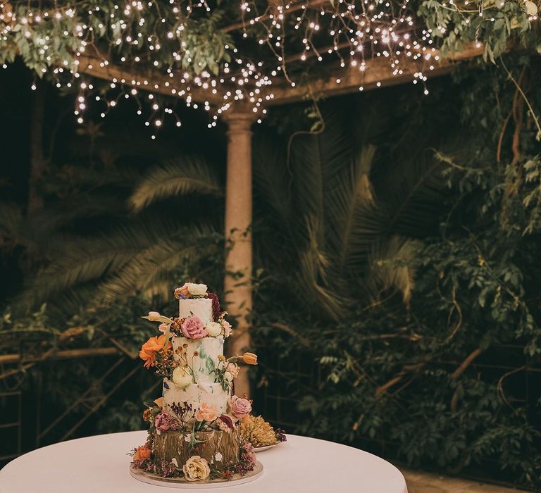 4 tier wedding cake with flower decor under a canopy of fairy lights 