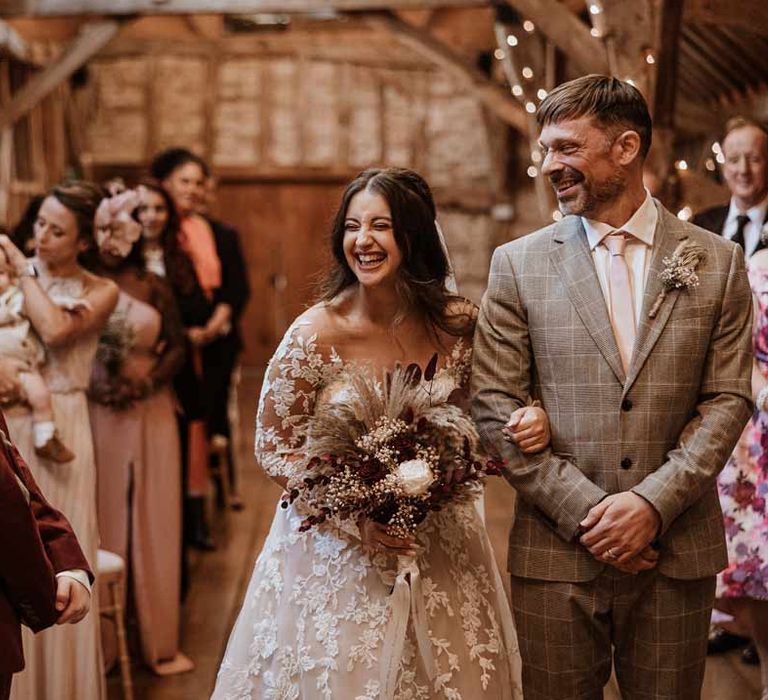 Bride in maroon bridal suit with grey waistcoat, dusky pink tie and autumnal dried flower boutonniere smiling at bride in long sleeve lace illusion sleeve wedding dress with overlay holding large autumnal boho dried flower bouquet 