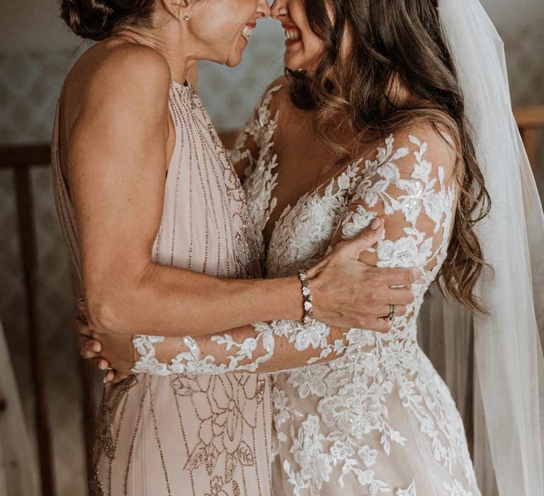 Bride in long sleeve lace illusion sleeve wedding dress with overlay hugging mother of the bride in dusky pink sleeveless dress and dusky pink fascinator 