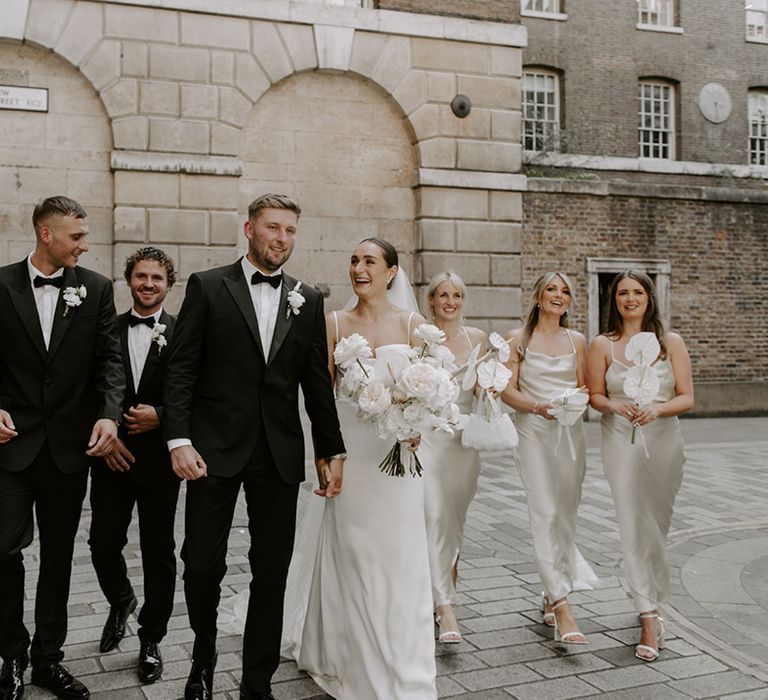 The wedding party with the groom and groomsmen in matching black tuxedos and the bride and bridesmaid in white wedding outfits 