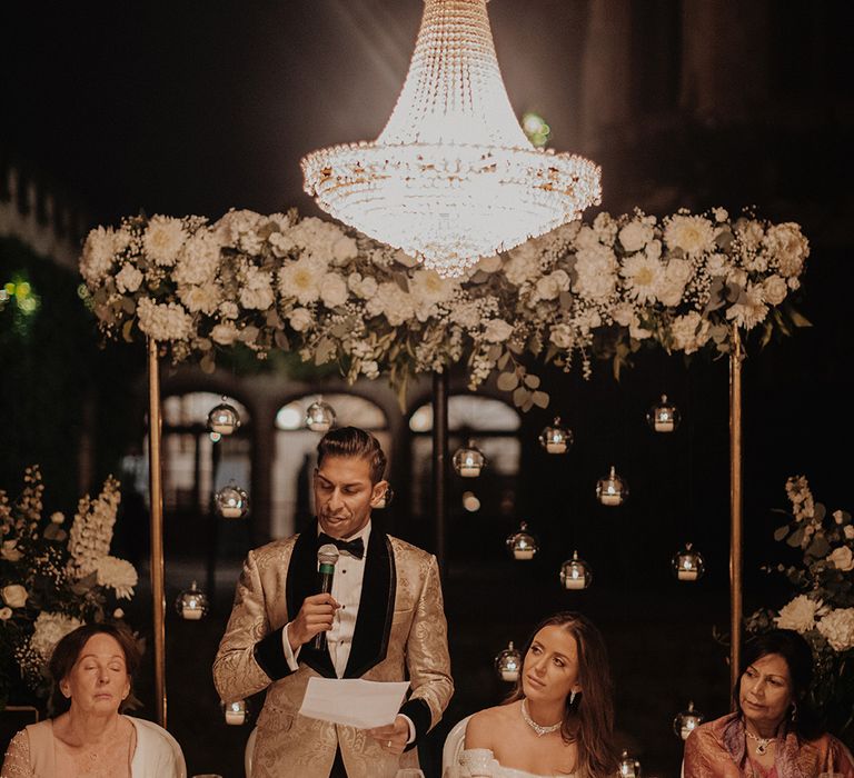 Groom gold blazer with black lining, black bowtie and black suit trousers giving wedding speech under wedding mandap with large bejewelled chandelier 