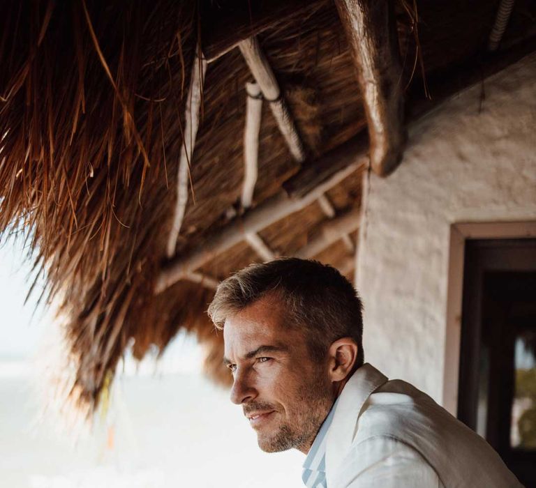 Groom standing on hotel balcony wearing mens white wedding suit