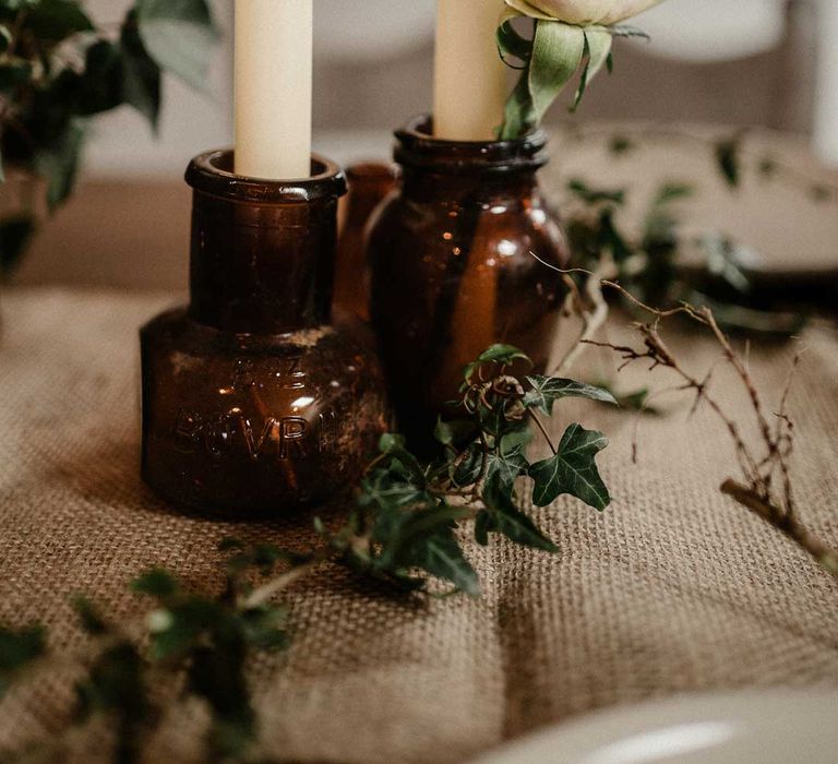 Beeswax dinner candles in small brown candle holder jars on burlap wedding table runner with ivy and dried flower decorations 