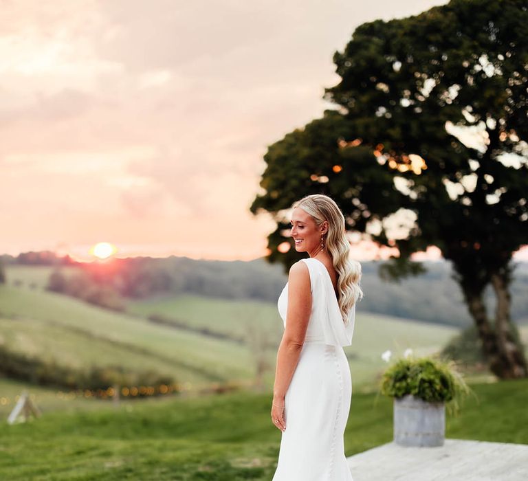 Bride in draped back Savin London wedding dress with train wearing her hair in Hollywood waves during the sunset 