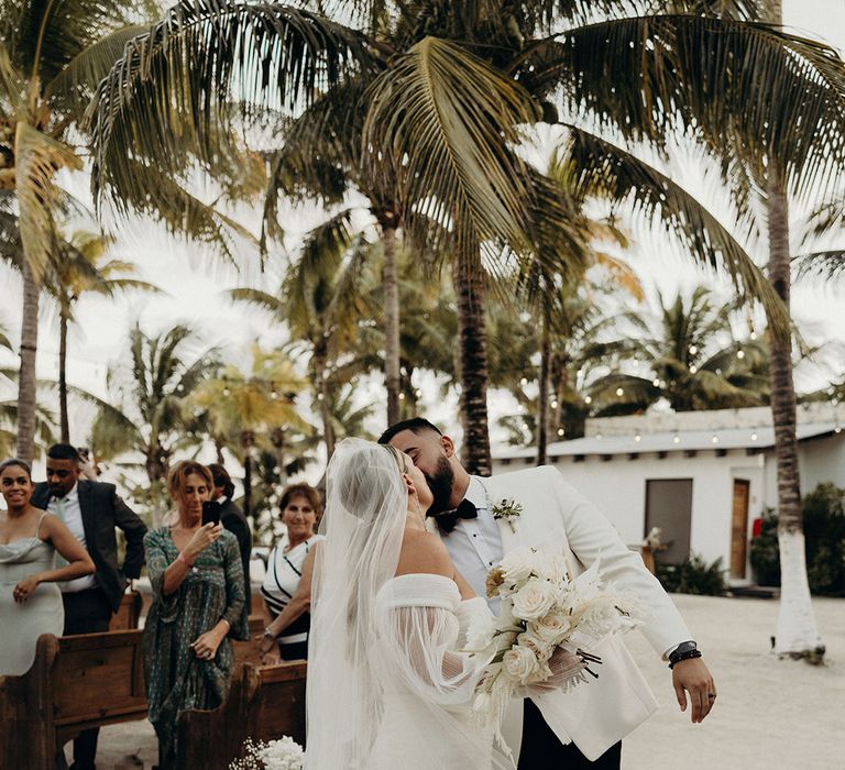 bride in a fishtail tulle wedding dress and veil kissing her groom in a white tuxedo jacket at the end of the aisle at their Blue Venado beach wedding 