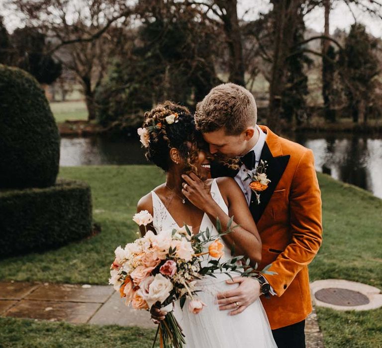 Groom in crushed burnt orange velvet grooms blazer, black bowtie and black suit trousers embracing bride in v-neck sleeveless wedding dress with 3D floral applique train holding blush and orange garden roses, poppies and carnations, dried flowers and eucalyptus wedding bouquet at Brinsop Court wedding venue